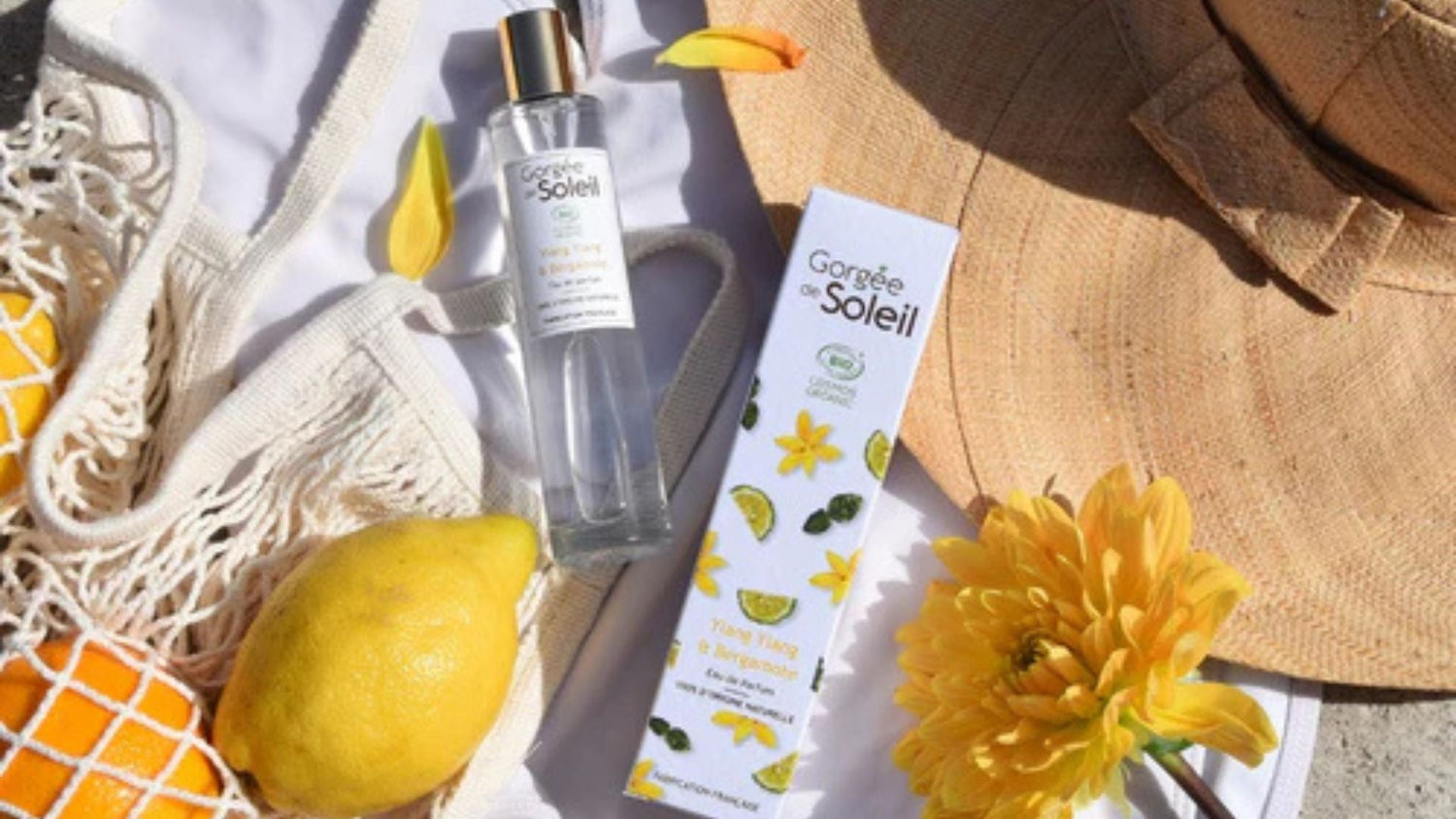 Eau de parfum bio "Gorgée de Soleil" aux notes d’Ylang-Ylang et de Bergamote, mise en scène estivale avec citron, fleur jaune, chapeau de paille et filet à provisions