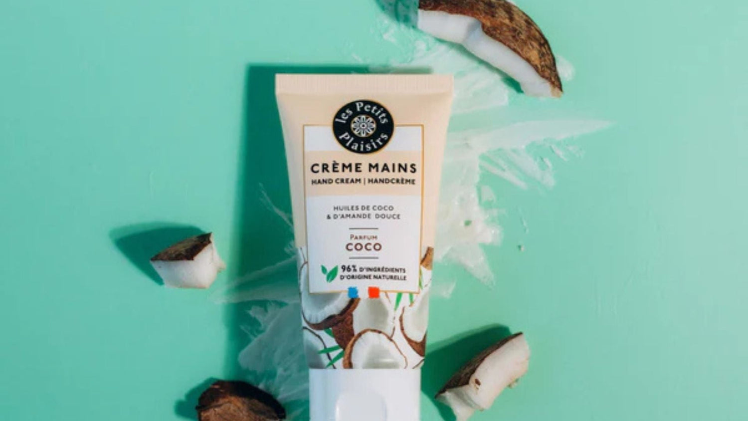 Crème mains parfum coco Les Petits Plaisirs à base d'huiles de coco et d'amande douce, posée sur un fond vert avec des morceaux de noix de coco