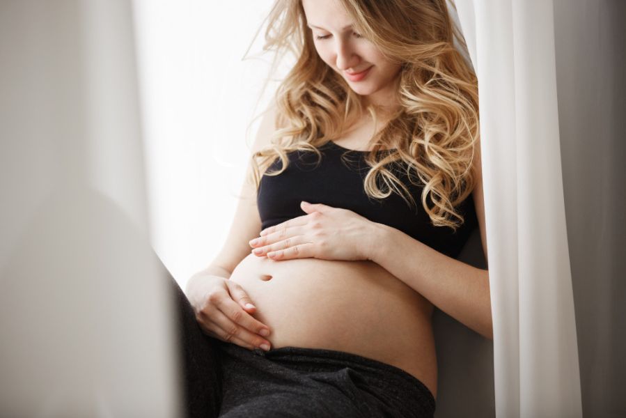 Femme enceinte souriante assise près d’une fenêtre, caressant son ventre avec douceur
